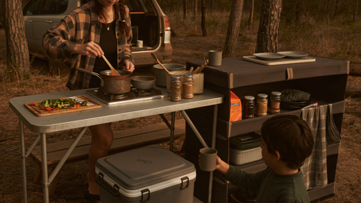Family camp kitchen setup at a forest campsite with folding table, two-burner stove, and tiered camp shelf at golden hour