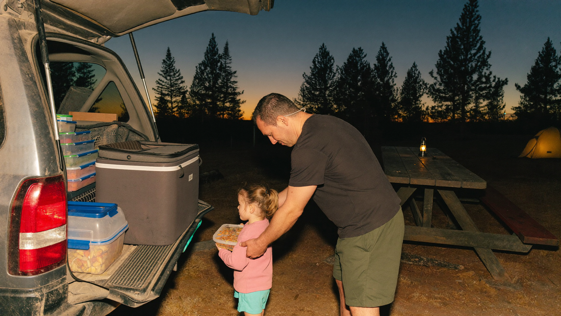 Father and daughter showing how to store food while camping by loading a soft cooler and sealed bins into their car at dusk
